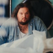 A man with dark hair and a beard wearing a white tshirt and denim shirt